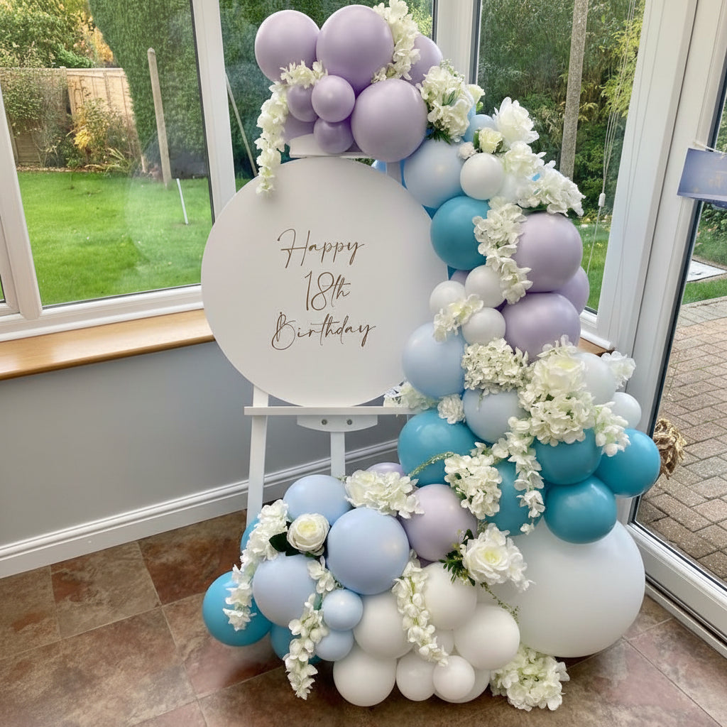 Panel de bienvenida con flores y globos azules, blancos y lavanda